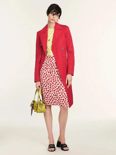 Model in a bright red coat and patterned skirt holding a lime green handbag against a seamless white studio background