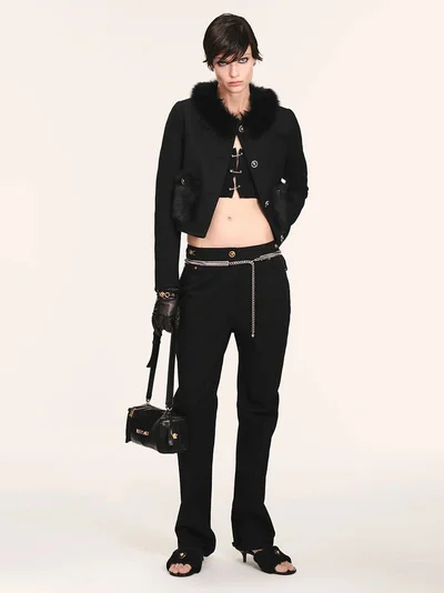 Model in black jacket with fur collar and trousers holding handbag against a seamless white background