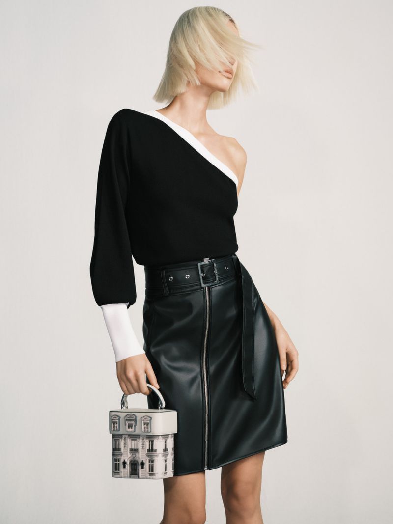 Fashion shot of a fair-skinned woman wearing a one-shoulder black top, a black leather skirt, and holding a small building-themed purse.