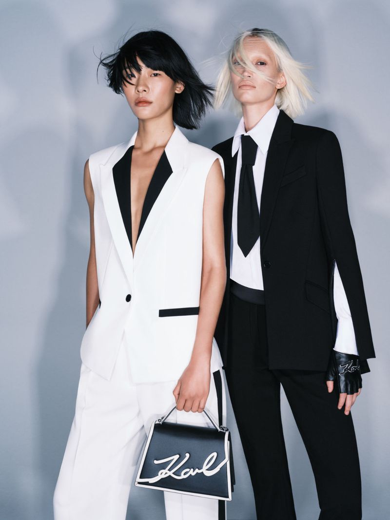 Two fashion models pose in studio wearing black and white suits with a black leather handbag with white signature lettering.