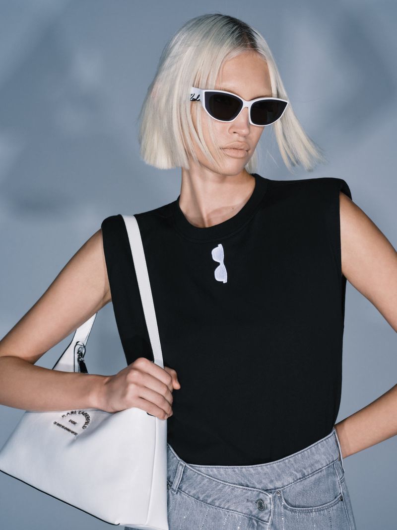 Adult woman poses wearing a black sleeveless top, light-wash jeans, and white sunglasses, holding a white tote bag in a photo studio.