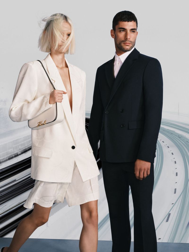 Two fashion models, a woman with a handbag and a man in a suit, pose against an abstract grey background in a studio shot.