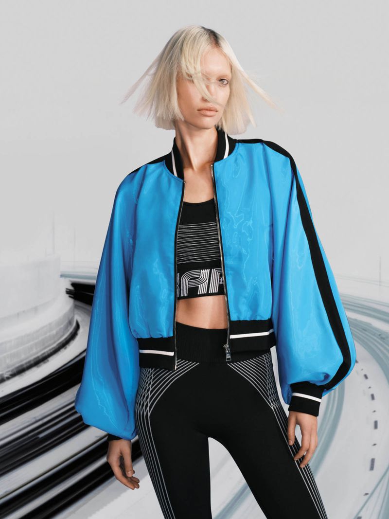Woman poses wearing a blue, black, and white bomber jacket with a matching black and white striped sports bra and leggings in a studio setting.