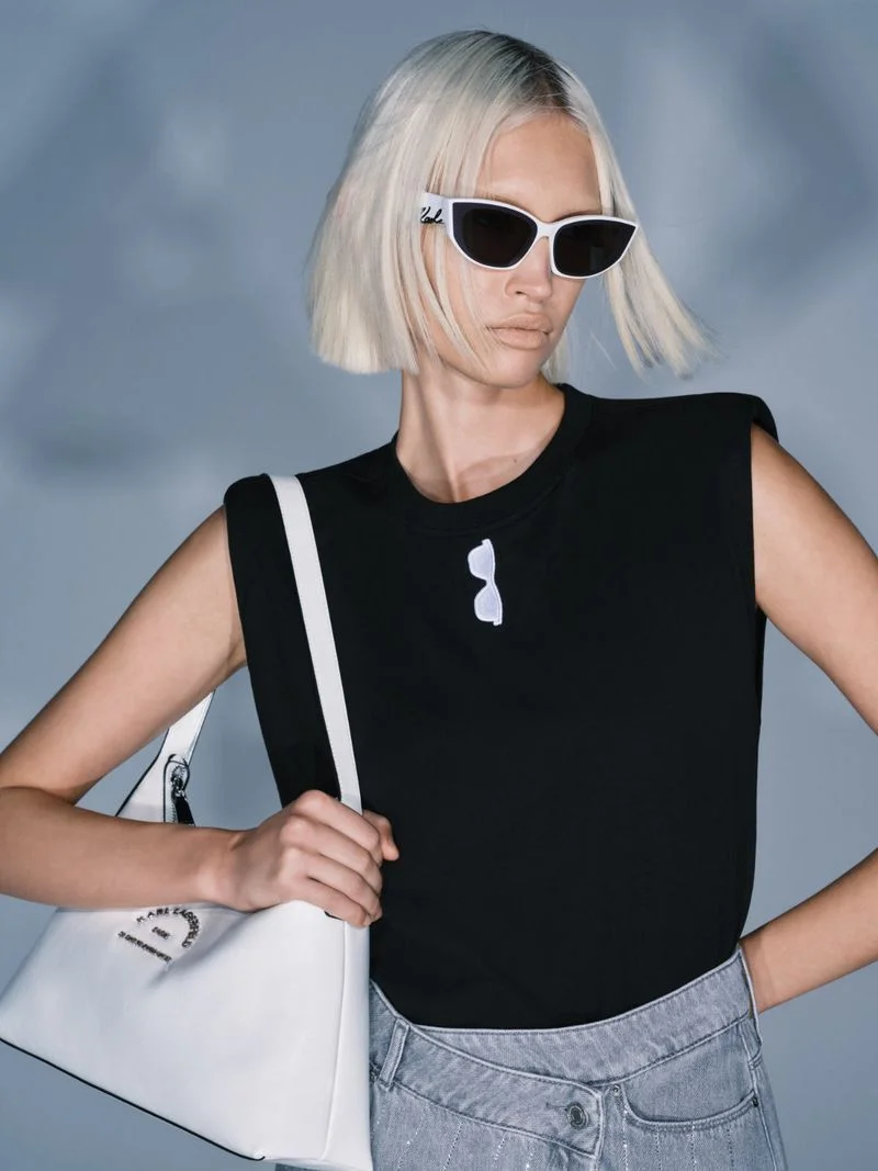 Patricia Zajac wears white cat-eye sunglasses, a black sleeveless top, and a white shoulder bag against a cool grey backdrop.