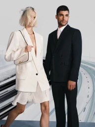 Patricia Zajac wearing an oversized white blazer and shorts beside a man in a black double-breasted suit against a highway backdrop.