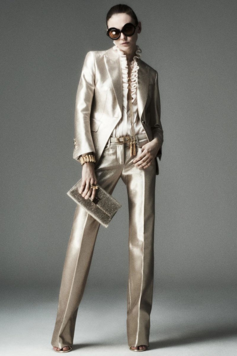 Model posing in a metallic champagne tailored pantsuit, ruffled blouse, oversized dark sunglasses, and holding a sparkling clutch.