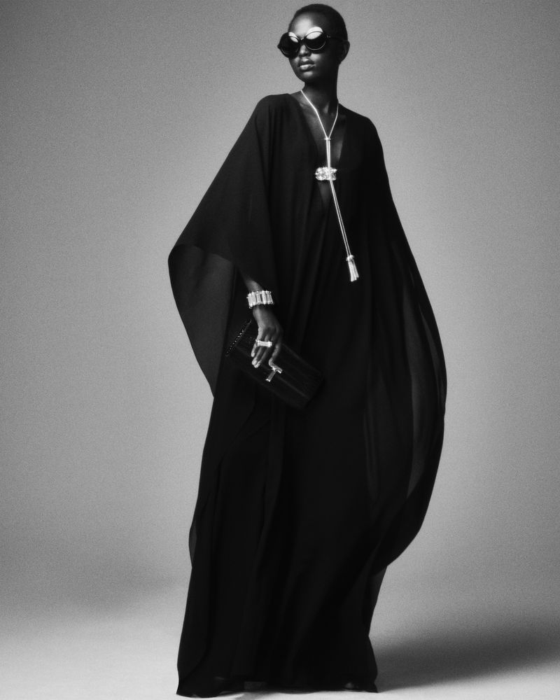 Black and white shot of a Black woman in a flowing black dress and sunglasses holding a clutch in a photo studio.