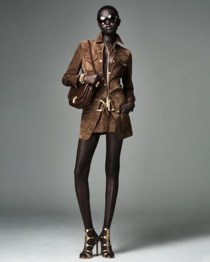 A dark-skinned female fashion model poses in brown suede jacket and skirt, sunglasses, hose, high heels, and a brown handbag on gray.