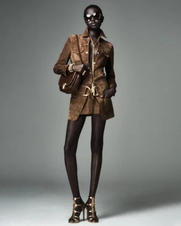 A dark-skinned female fashion model poses in brown suede jacket and skirt, sunglasses, hose, high heels, and a brown handbag on gray.