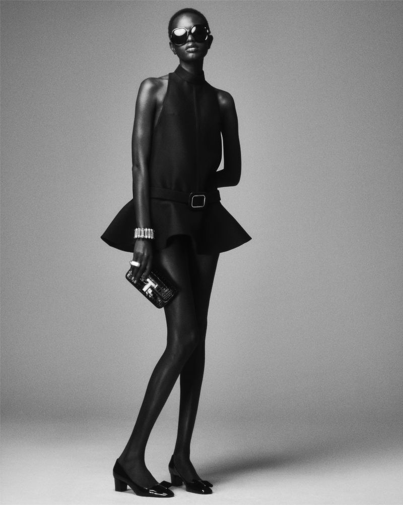 Black and white fashion shot of a slender woman with sunglasses, wearing a minidress, bracelet, and heels, and holding a clutch purse.