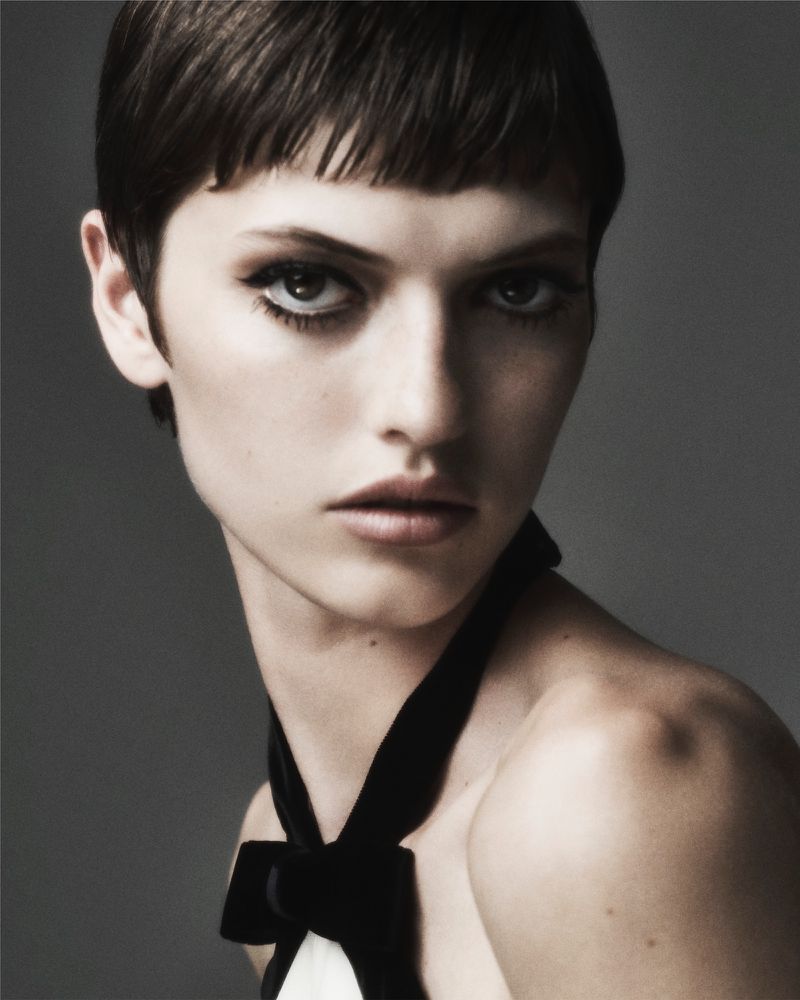 Close-up portrait of a model with a short, dark haircut, striking eye makeup, and a black velvet bow, set against a grey backdrop.