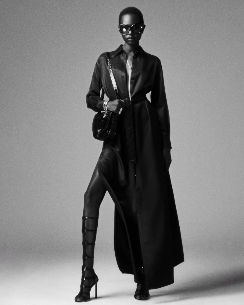 A single Black woman models a chic black belted coat dress with gladiator sandals and sunglasses against a neutral gradient backdrop.