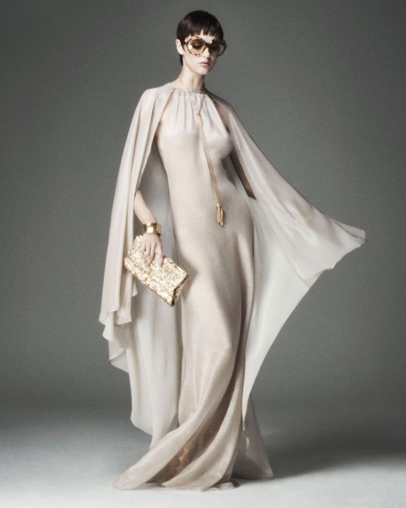Elegant woman in flowing beige gown with cape, gold accessories, and sunglasses, posing against a neutral backdrop in a fashion-forward style.