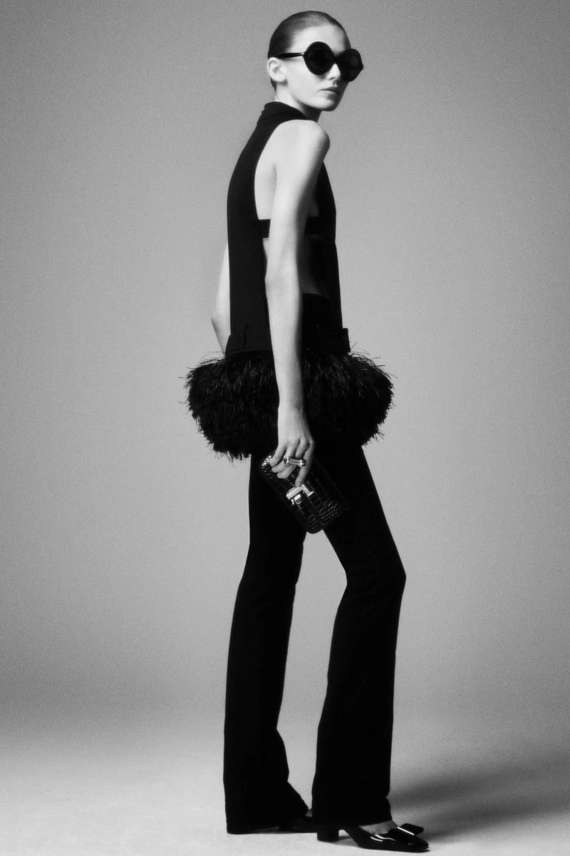 Black-and-white full shot of a woman in a dark sleeveless outfit with feather details, flared trousers, and heart-shaped sunglasses.