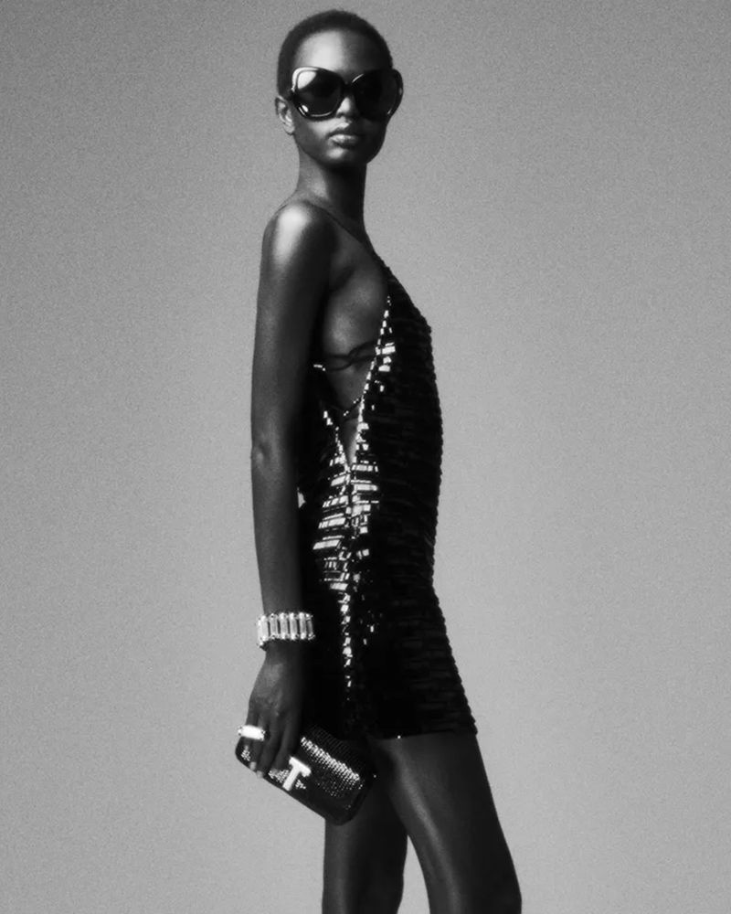 Model wearing a black sequined mini dress, oversized dark sunglasses, and holding a black clutch against a grey studio backdrop.