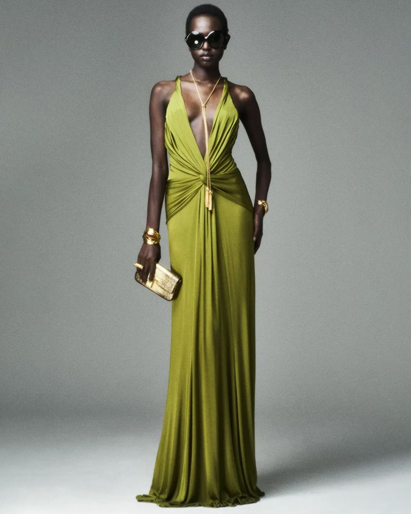 A dark-skinned female model poses in a studio, wearing a flowing chartreuse dress with sunglasses and gold jewelry, holding a gold clutch.