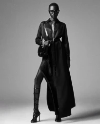 Model Nyaduola Gabriel poses in a long black shirt dress, oversized sunglasses, and caged stiletto heels against a grey studio backdrop.