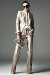 Model posing in a metallic champagne tailored pantsuit, ruffled blouse, oversized dark sunglasses, and holding a sparkling clutch.