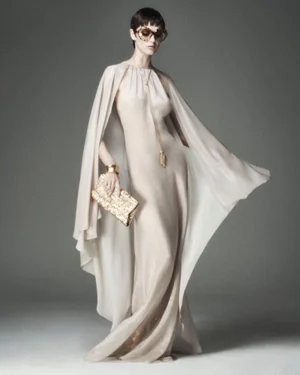 Model in a flowing cape dress wearing oversized sunglasses and holding a gold clutch against a grey seamless backdrop
