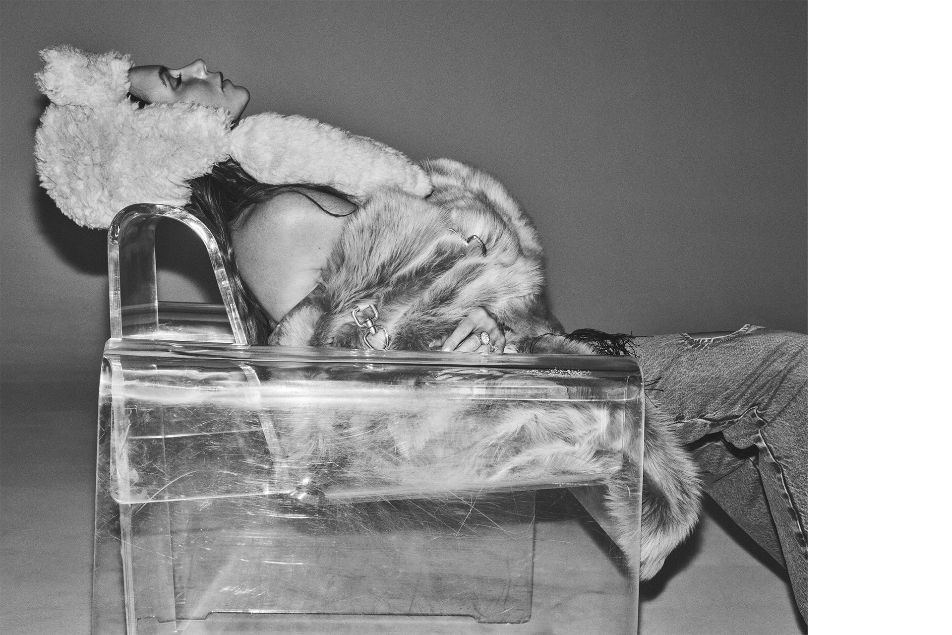 Monochrome shot of a woman in a fur coat and hat, reclining in a transparent chair against a gray background, contemplative and stylish.
