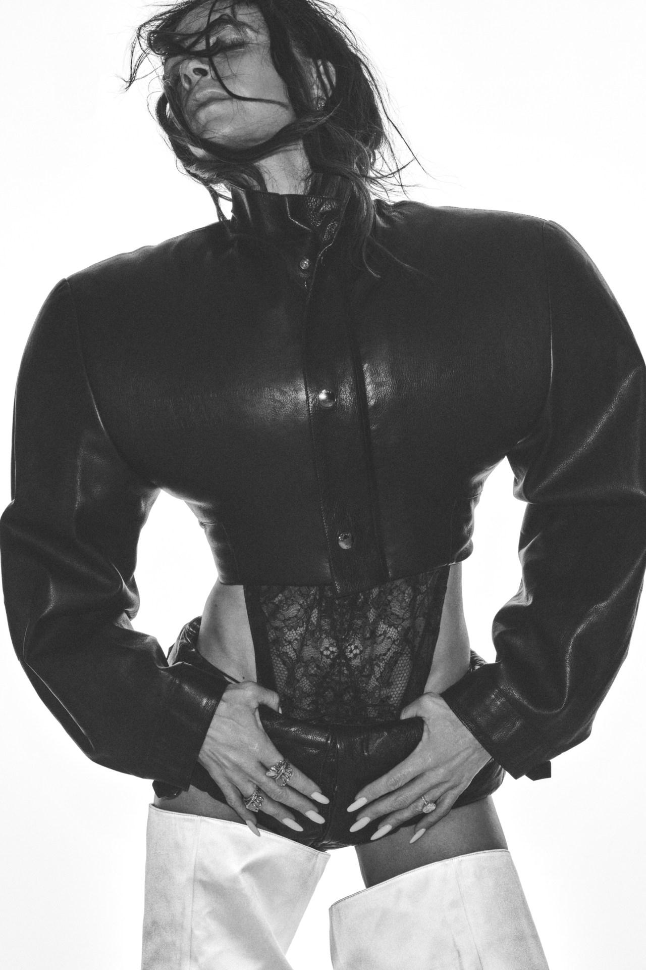 Monochrome fashion shot: Woman in leather jacket and lace bodysuit, hands on hips, against a white background, for editorial style.
