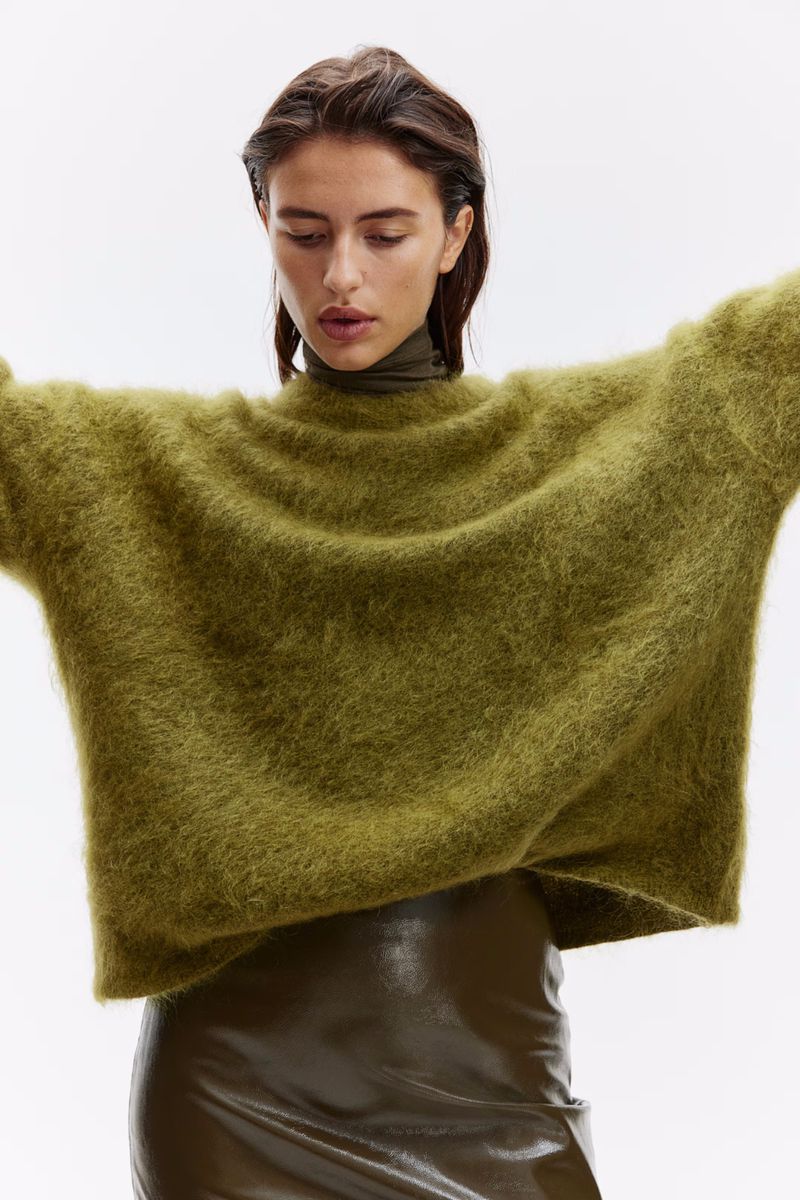 A woman models an oversized, shaggy olive-green sweater with a turtle neck and a shiny olive-green mini skirt against a white background.