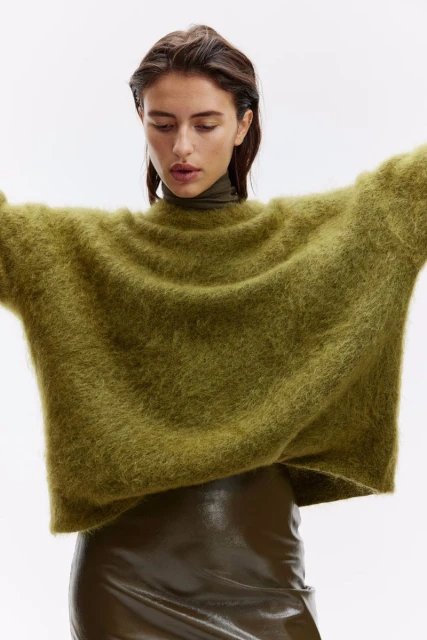 A woman models an oversized, shaggy olive-green sweater with a turtle neck and a shiny olive-green mini skirt against a white background.