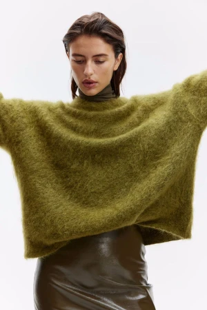 A woman models an oversized, shaggy olive-green sweater with a turtle neck and a shiny olive-green mini skirt against a white background.