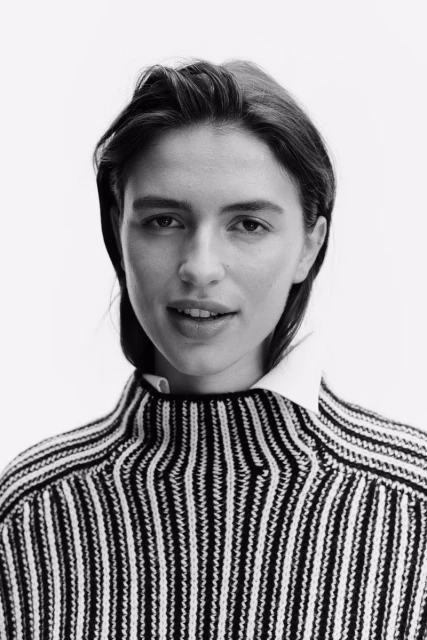 Headshot of a young woman with short brown hair wearing a black and white striped sweater with a white collared shirt underneath.