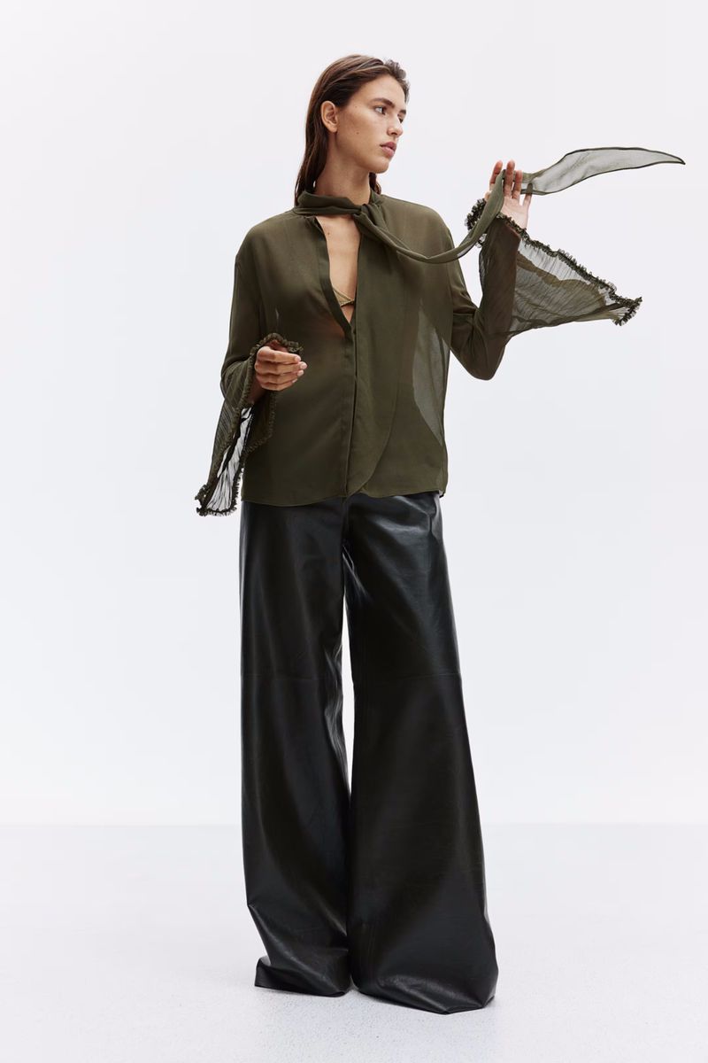 Full shot of a female model in an olive-green blouse with a sheer scarf and leather trousers against a white backdrop in a photo studio.