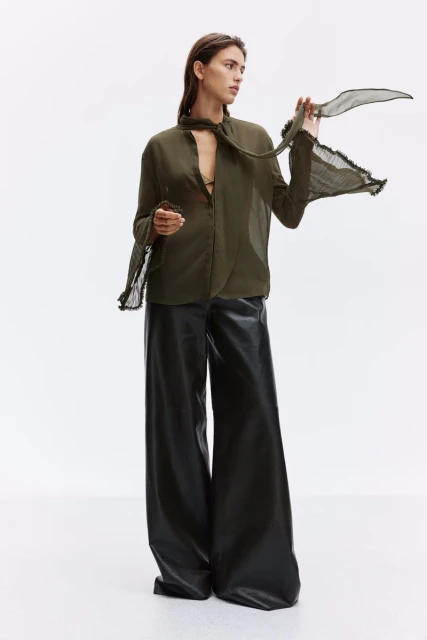 Full shot of a female model in an olive-green blouse with a sheer scarf and leather trousers against a white backdrop in a photo studio.