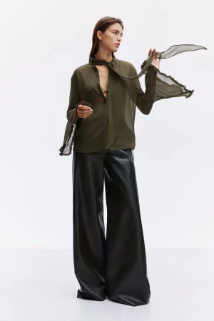 Full shot of a female model in an olive-green blouse with a sheer scarf and leather trousers against a white backdrop in a photo studio.