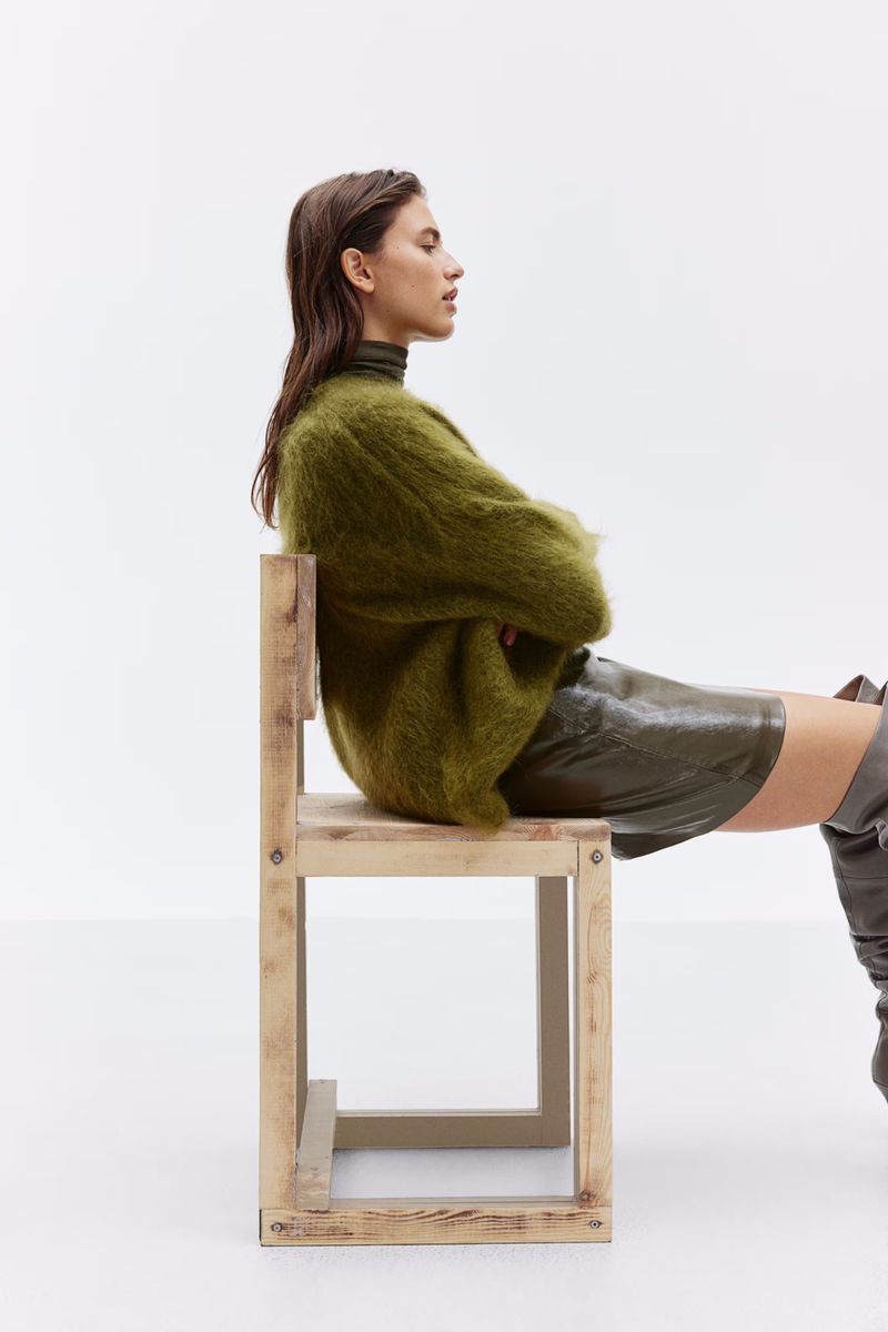 Model seated on a wooden chair wearing an olive green mohair sweater and knee-high boots against a white seamless background