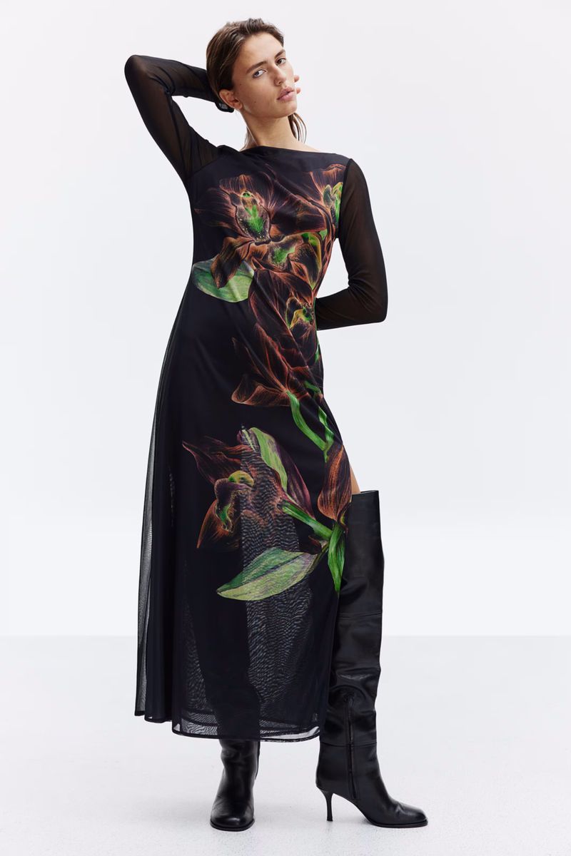 Model in a long black mesh dress with botanical print and black boots against a white studio background.