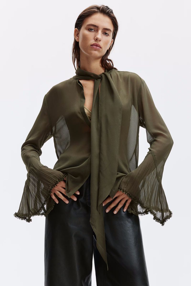 Fashion shot of a woman in a sheer olive blouse with a bow tie, black pants, against a bright white background in a studio setting.