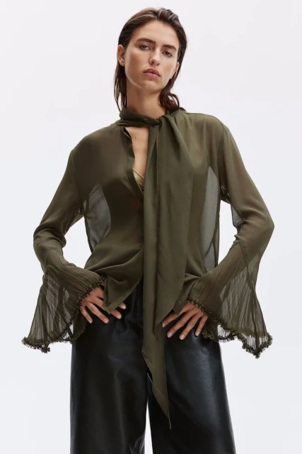 Fashion shot of a woman in a sheer olive blouse with a bow tie, black pants, against a bright white background in a studio setting.