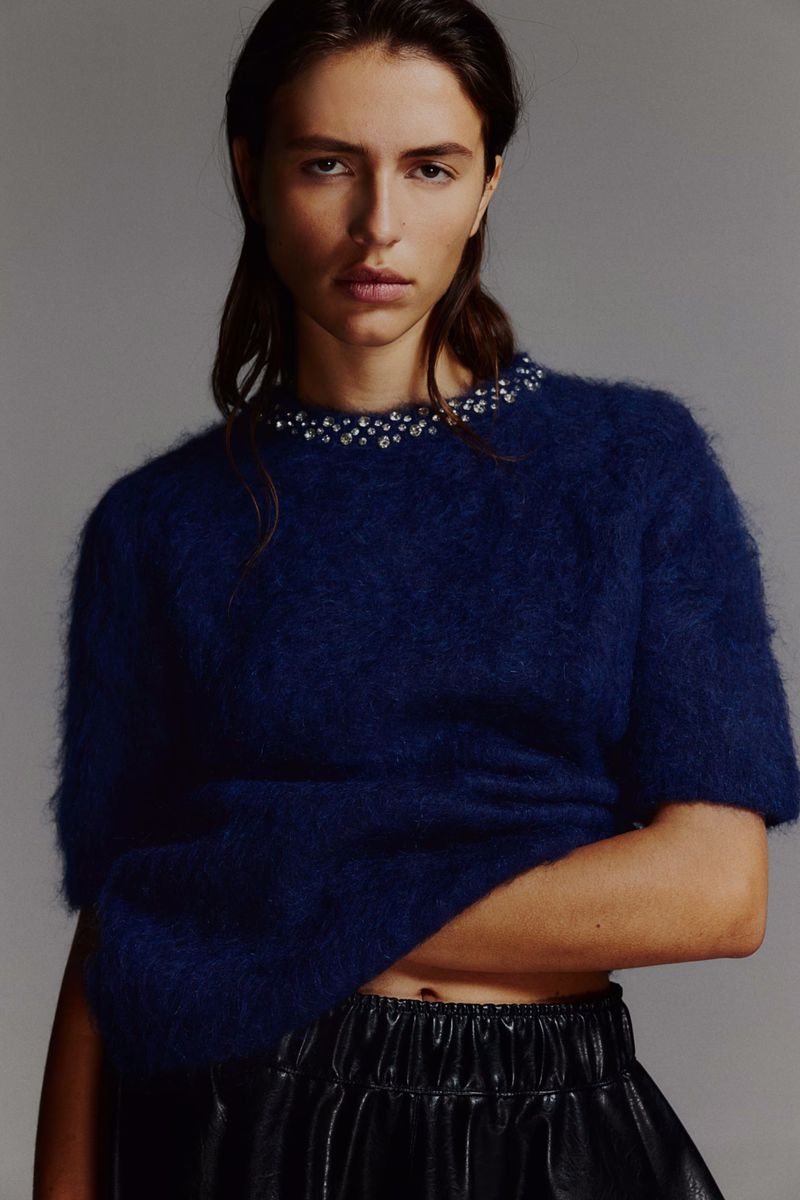 Woman in a fashion shot wearing a blue sweater with embellishments and a black skirt, photographed in a studio with neutral background.