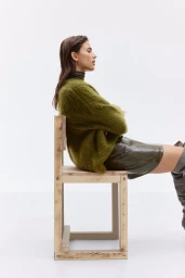 Model seated on a wooden chair wearing an olive green mohair sweater and knee-high boots against a white seamless background