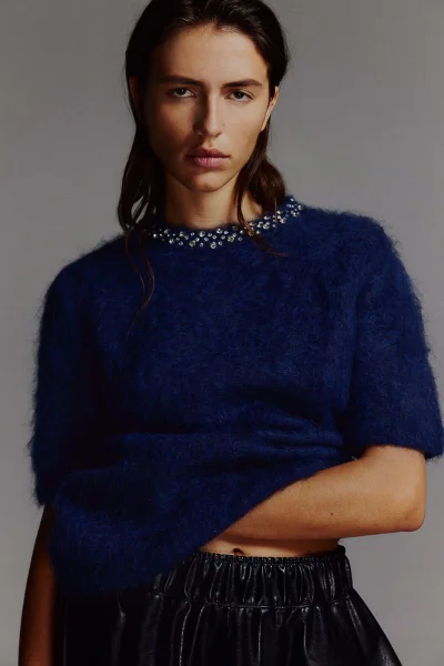 Model in a dark blue textured sweater with a beaded neckline posing against a neutral grey studio backdrop.