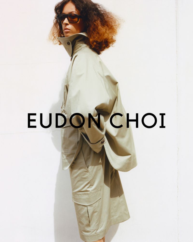 Eudon Choi fashion advertisement featuring a woman in a light-green jacket and shorts, posing against a bright white background in studio lighting.