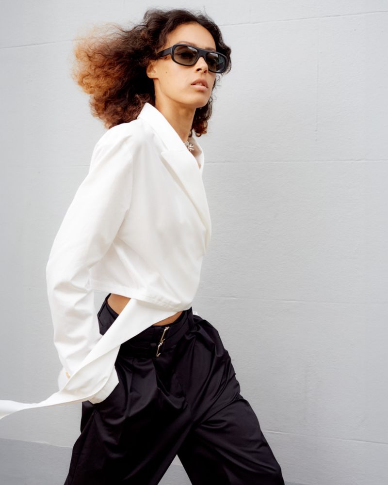 An adult female fashion model with curly hair wears sunglasses, a white top, and black satin pants while posing against a grey wall.