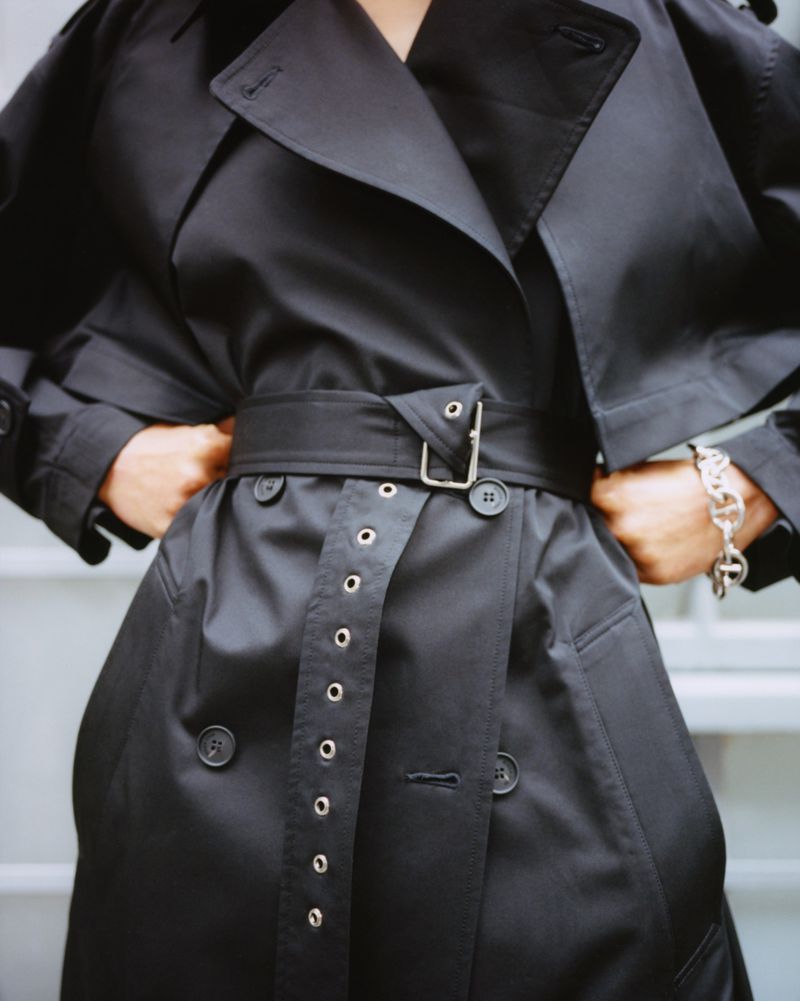 Close-up on the torso of a person wearing a black trench coat with a belt and silver accents, accessorized with a silver bracelet.