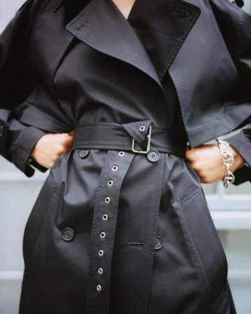 Close-up on the torso of a person wearing a black trench coat with a belt and silver accents, accessorized with a silver bracelet.