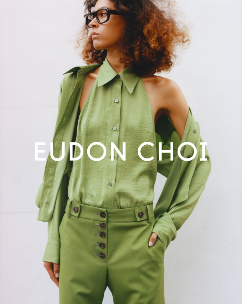 Fashion shot of a curly-haired woman wearing a green two-piece outfit and glasses against a white wall, showcasing contemporary style.