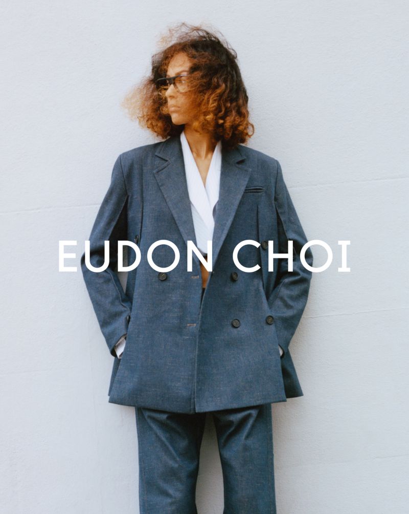 An adult woman wearing sunglasses poses in a relaxed blue suit in front of a white wall in a fashion-editorial style outdoor shot.