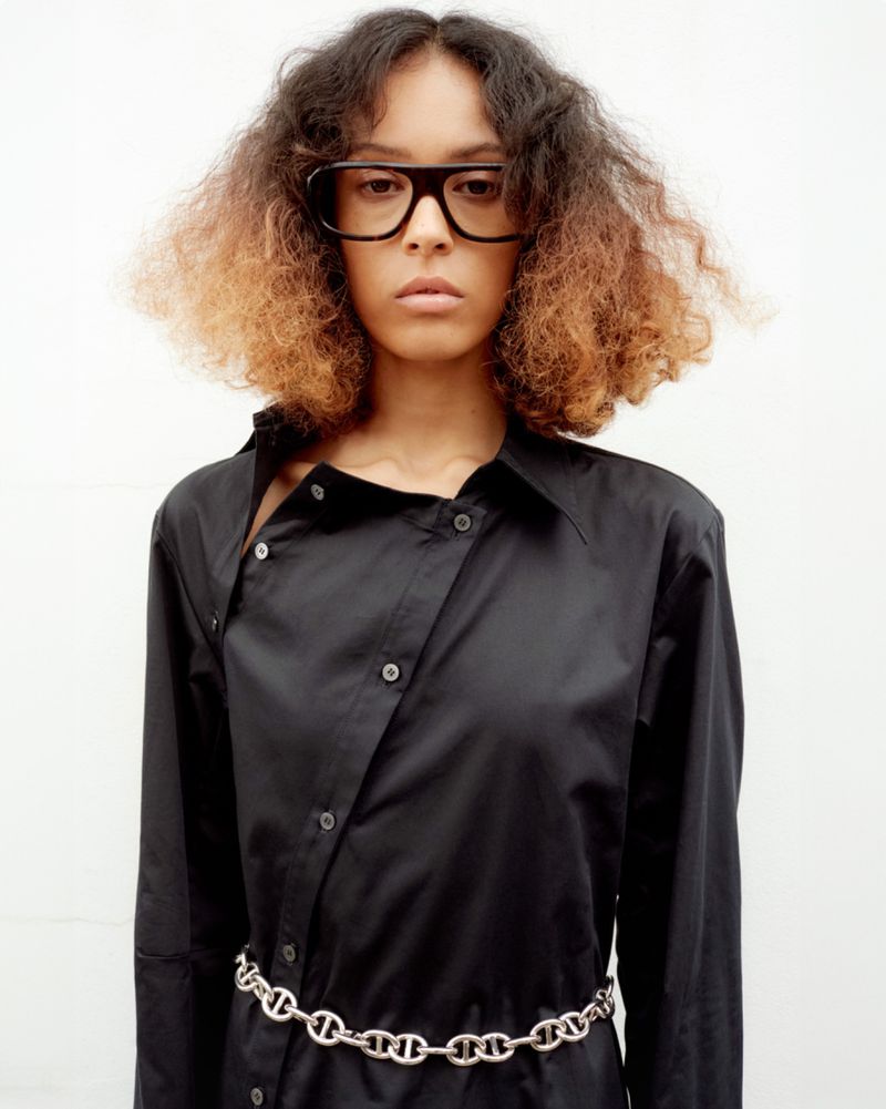 Portrait of a woman with curly hair wearing glasses and a black shirt dress with a silver chain belt in front of a white background.
