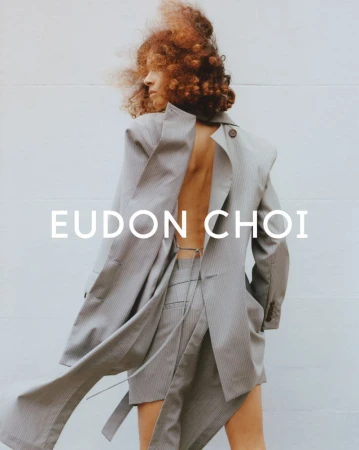 Fashion shot of a woman with curly red hair wearing a striped blazer dress in front of a white wall with the text Eudon Choi.