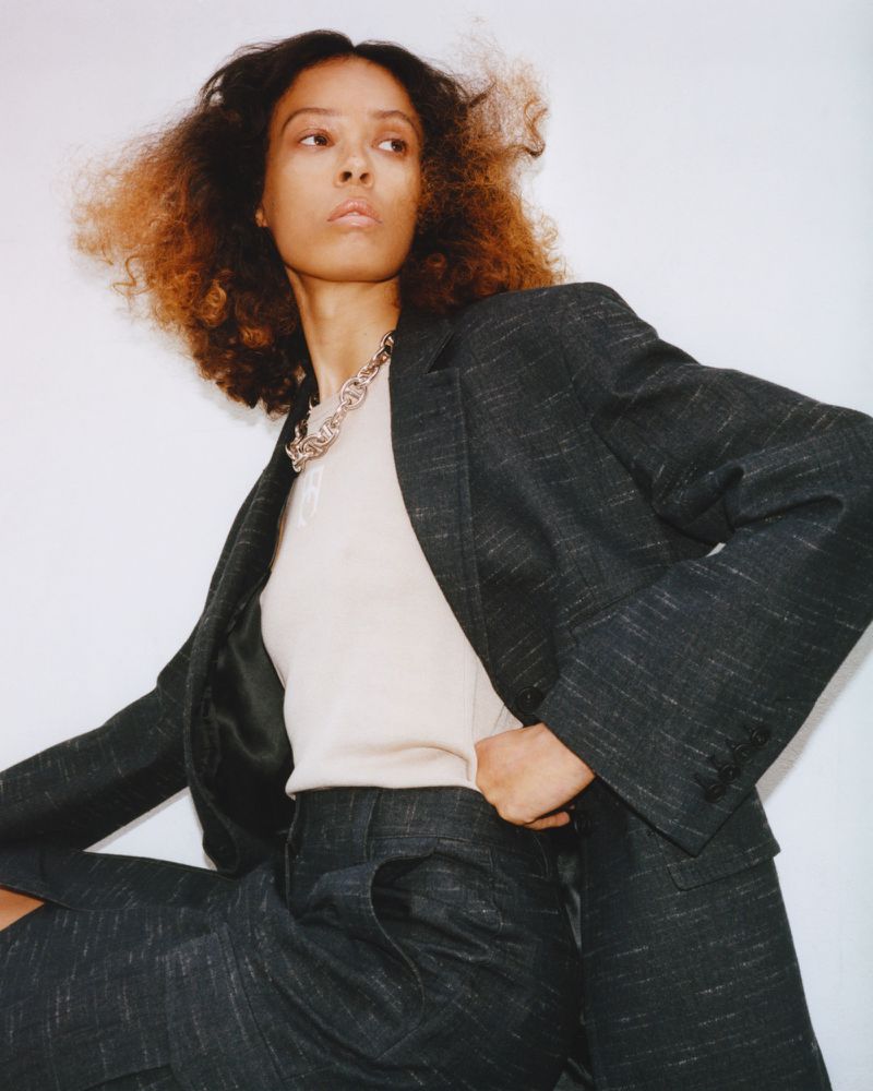A model poses in a dark grey suit with a tan sweater, seated against a white background in a studio environment for a fashion editorial.