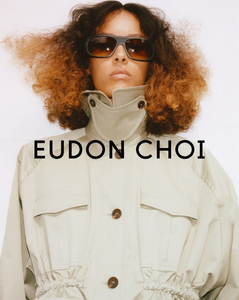 Fashion portrait of a woman wearing sunglasses and a beige jacket, posing against a white backdrop with the Eudon Choi logo.