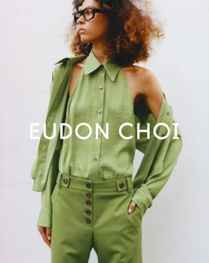 Model wearing a green sleeveless shirt and matching trousers with dark framed glasses against a white background